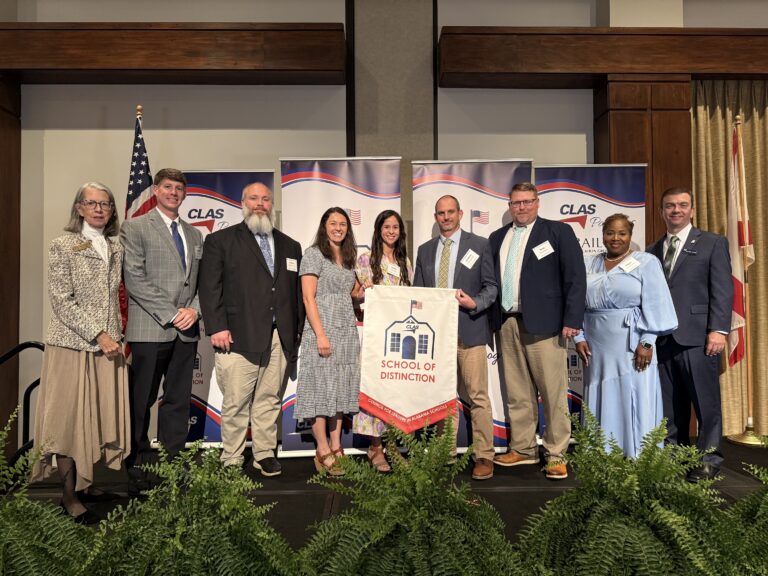 Group of nine educators and school leaders stand on stage at the CLAS School of Distinction Awards ceremony, posing with a School of Distinction banner. American and Alabama flags frame the stage, with CLAS sponsor backdrops behind them and green ferns lining the front of the platform.