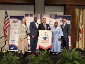 Group of five educators and school leaders stand on stage at the CLAS School of Distinction Awards ceremony, posing with a School of Distinction banner. American and Alabama flags frame the stage, with CLAS sponsor backdrops behind them and green ferns lining the front of the platform.