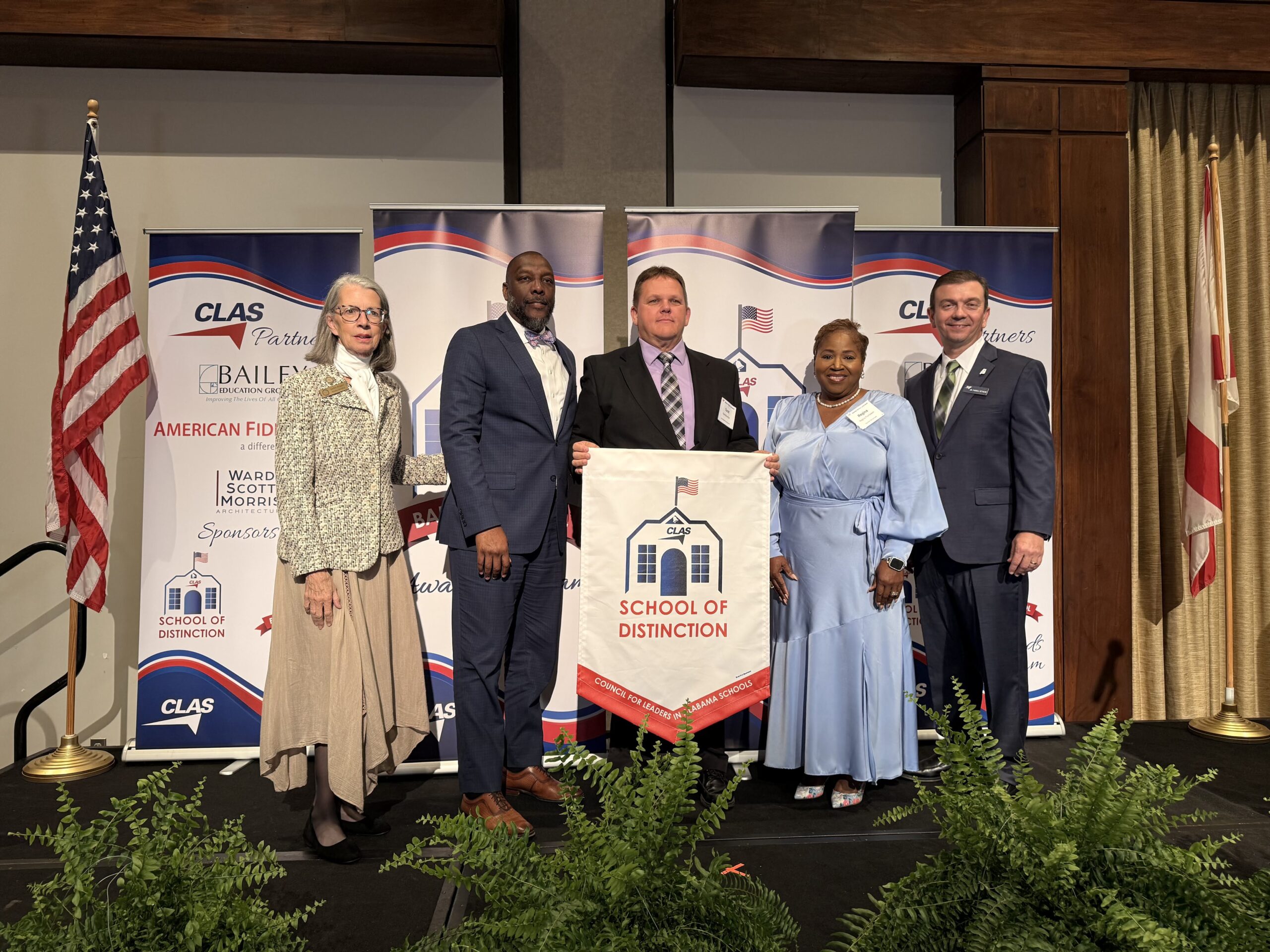 Group of five educators and school leaders stand on stage at the CLAS School of Distinction Awards ceremony, posing with a School of Distinction banner. American and Alabama flags frame the stage, with CLAS sponsor backdrops behind them and green ferns lining the front of the platform.