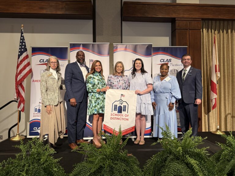 Group of seven educators and school leaders stand on stage at the CLAS School of Distinction Awards ceremony, posing with a School of Distinction banner. American and Alabama flags frame the stage, with CLAS sponsor backdrops behind them and green ferns lining the front of the platform.