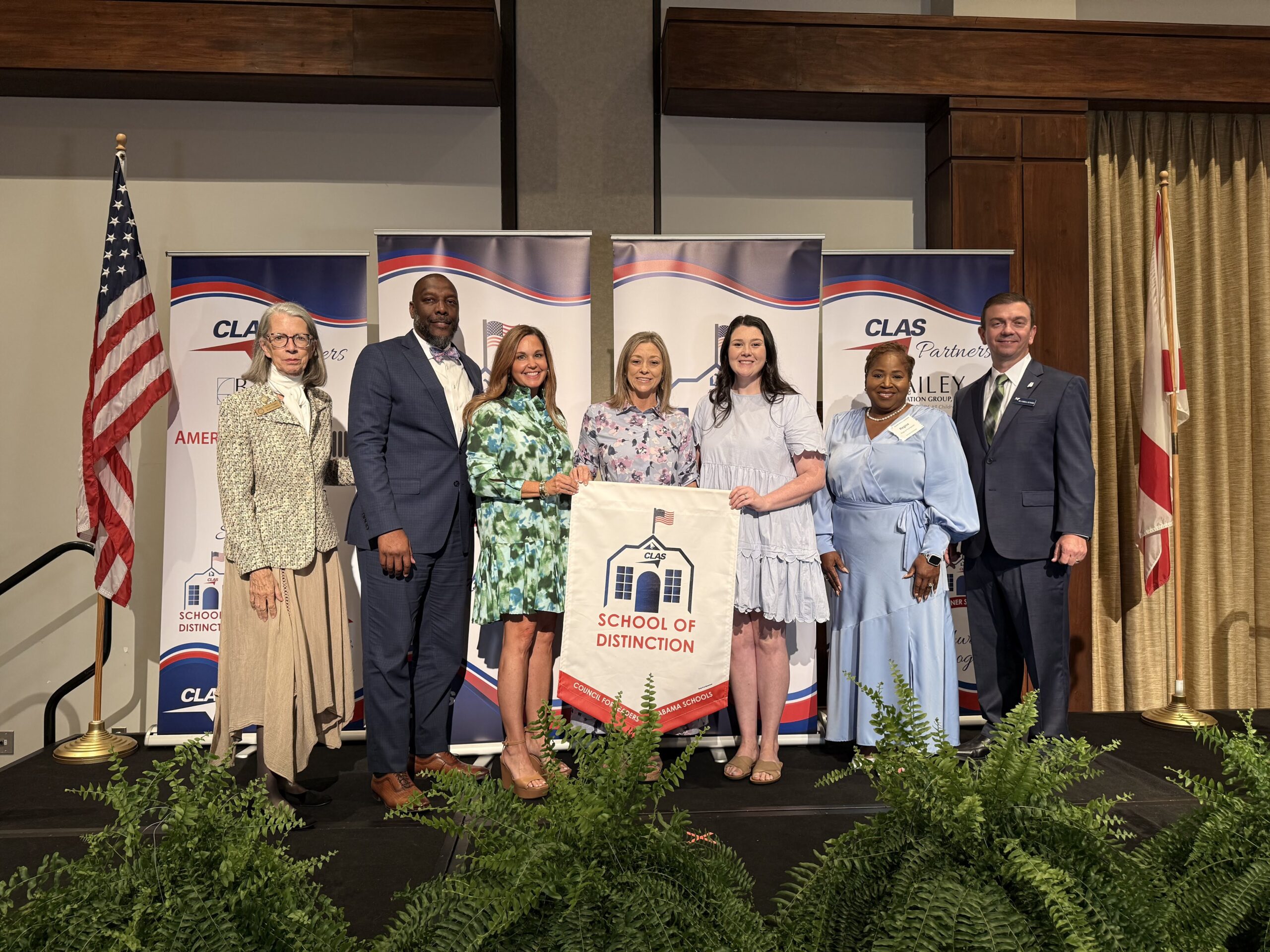 Group of seven educators and school leaders stand on stage at the CLAS School of Distinction Awards ceremony, posing with a School of Distinction banner. American and Alabama flags frame the stage, with CLAS sponsor backdrops behind them and green ferns lining the front of the platform.