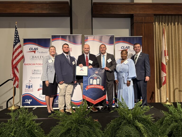 Group of seven educators and school leaders stand on stage at the CLAS Banner School Awards ceremony, posing with a Banner School banner and award plaque. American and Alabama flags frame the stage, with CLAS sponsor backdrops behind them and green ferns lining the front of the platform.