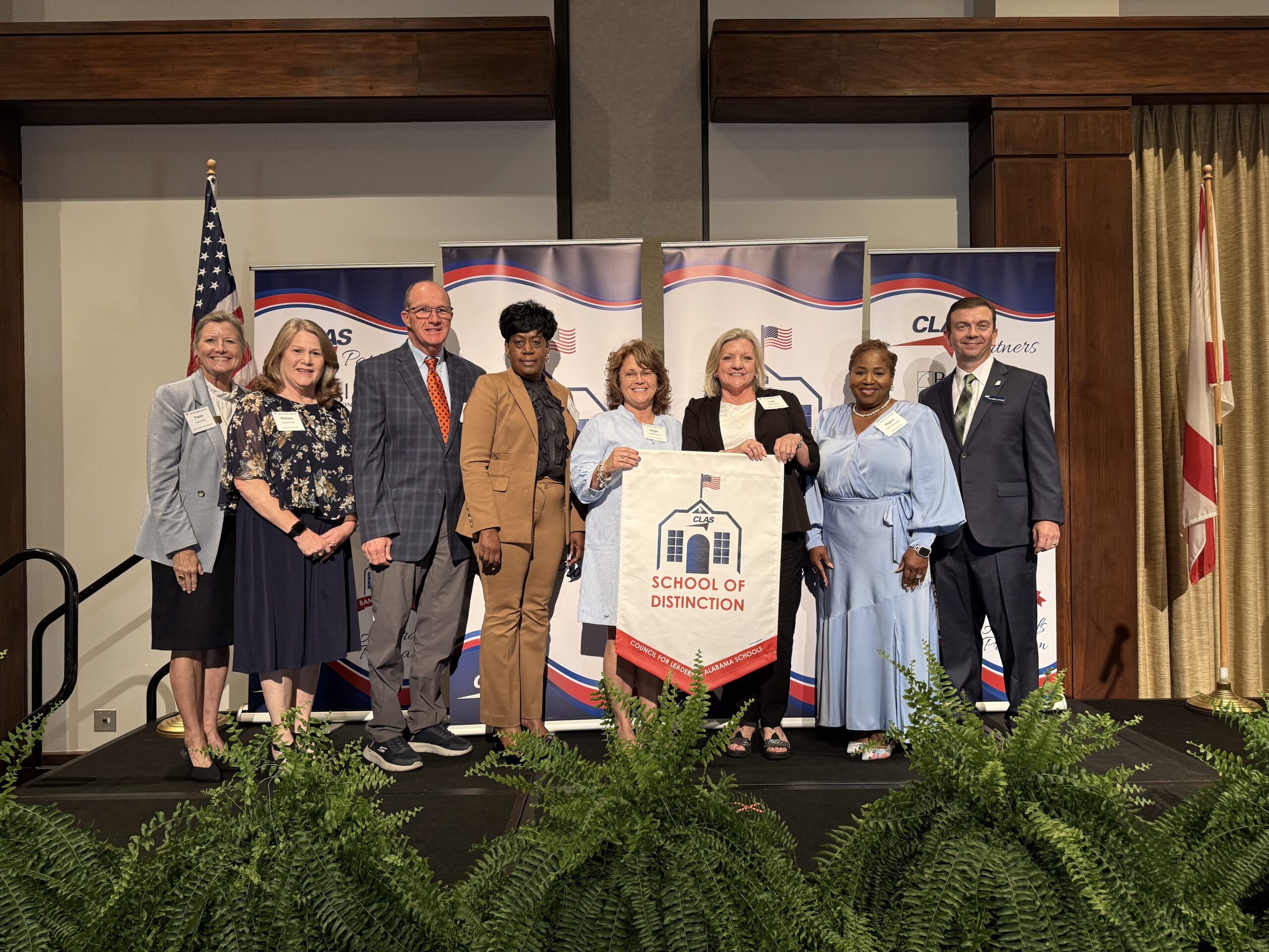 Group of eight educators and school leaders stand on stage at the CLAS School of Distinction Awards ceremony, posing with a School of Distinction banner. American and Alabama flags frame the stage, with CLAS sponsor backdrops behind them and green ferns lining the front of the platform.