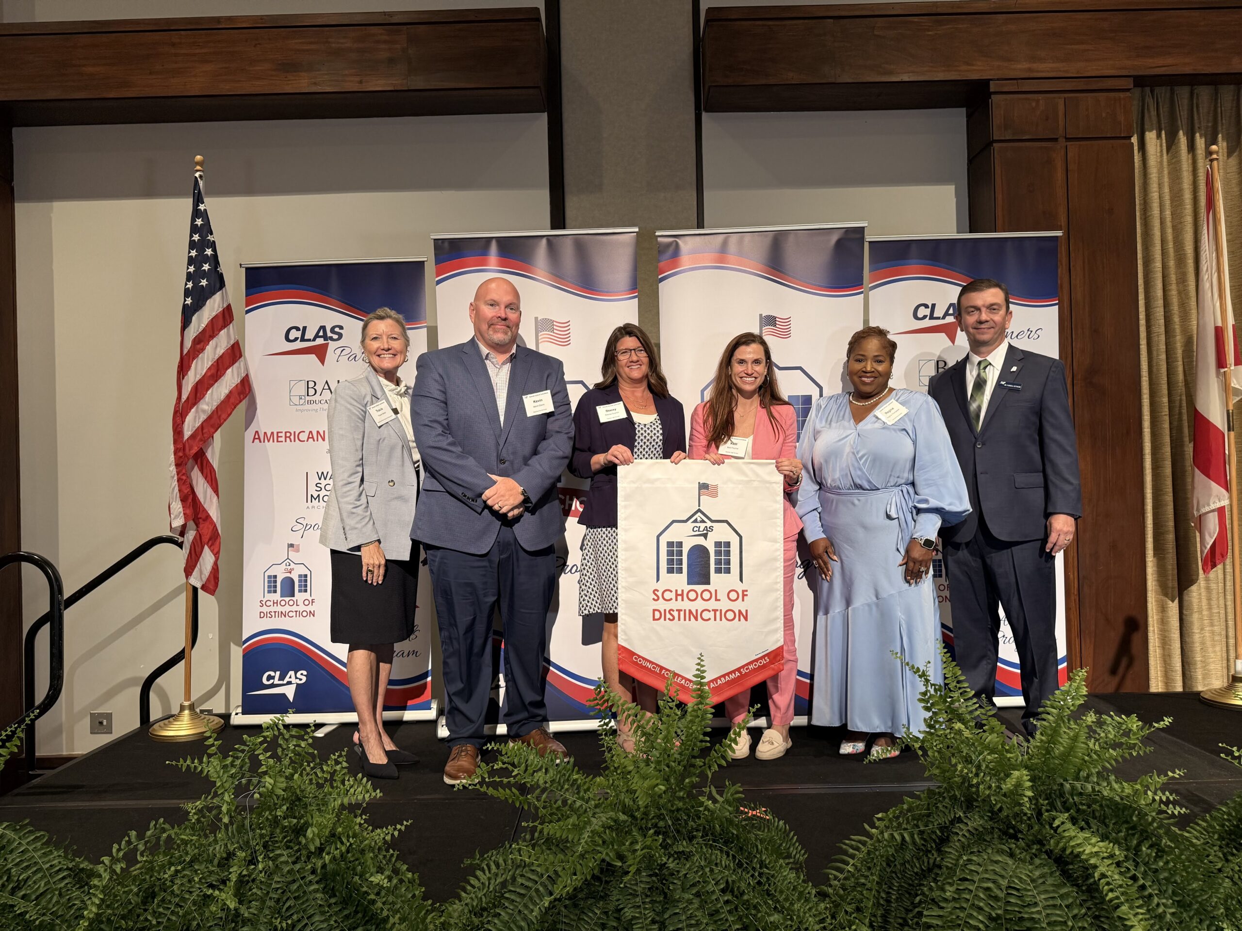 Group of seven educators and school leaders stand on stage at the CLAS School of Distinction Awards ceremony, posing with a School of Distinction banner. American and Alabama flags frame the stage, with CLAS sponsor backdrops behind them and green ferns lining the front of the platform.