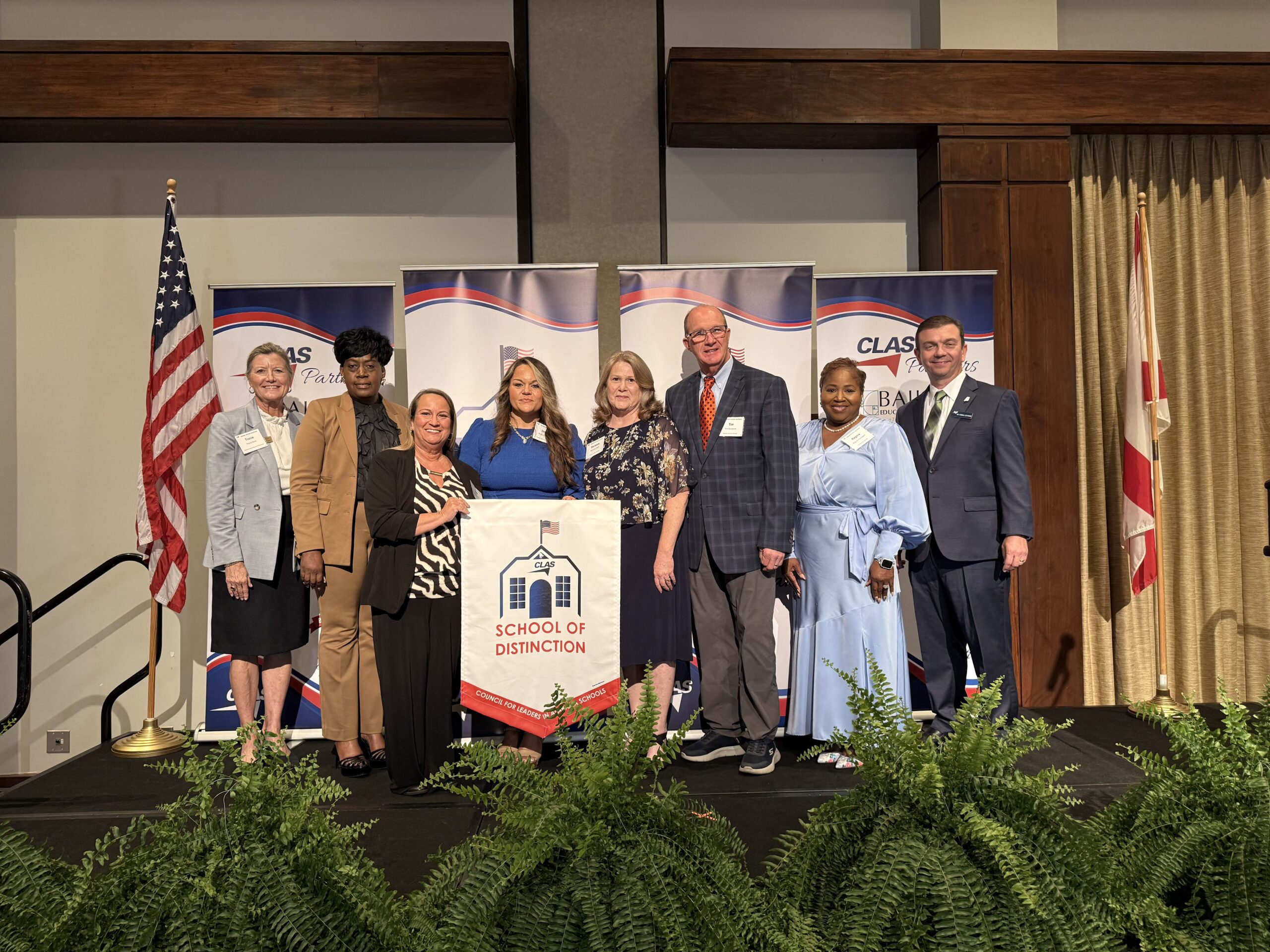 Group of eight educators and school leaders stand on stage at the CLAS School of Distinction Awards ceremony, posing with a School of Distinction banner. American and Alabama flags frame the stage, with CLAS sponsor backdrops behind them and green ferns lining the front of the platform.