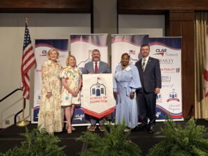 Five educators and school leaders stand on stage at the CLAS School of Distinction Awards ceremony, posing with a School of Distinction banner. American and Alabama flags frame the stage, with CLAS sponsor backdrops behind them and green ferns lining the front of the platform.
