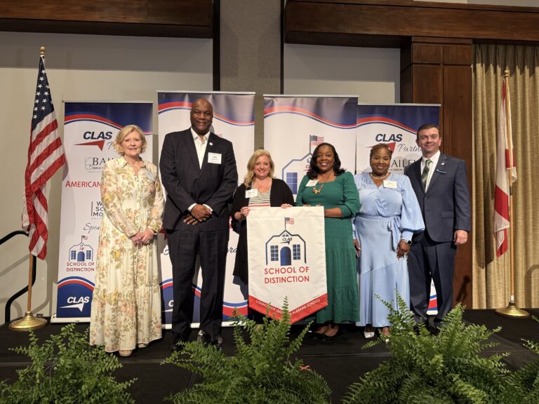 Group of six educators and school leaders stand on stage at the CLAS School of Distinction Awards ceremony, posing with a School of Distinction banner. American and Alabama flags frame the stage, with CLAS sponsor backdrops behind them and green ferns lining the front of the platform.