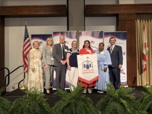 Group of seven educators and school leaders stand on stage at the CLAS School of Distinction Awards ceremony, posing with a School of Distinction banner. American and Alabama flags stand on either side of the stage, with CLAS sponsor backdrops and green ferns lining the front of the platform.