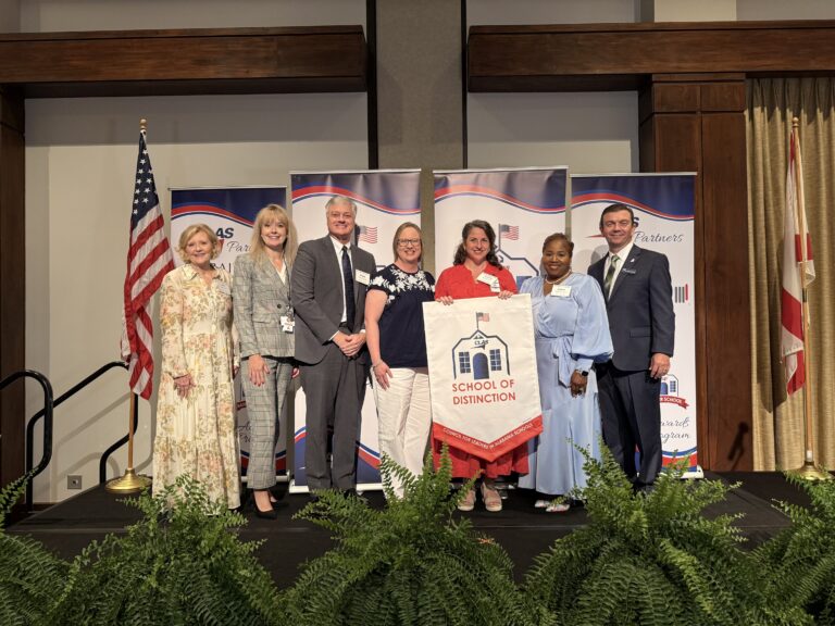Group of seven educators and school leaders stand on stage at the CLAS School of Distinction Awards ceremony, posing with a School of Distinction banner. American and Alabama flags stand on either side of the stage, with CLAS sponsor backdrops and green ferns lining the front of the platform.