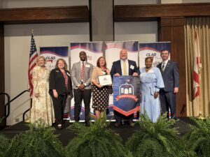 Group of seven educators and school leaders stand on stage at the CLAS Banner School Awards ceremony, posing with a Banner School banner and award plaque. American and Alabama flags flank the stage, with CLAS sponsor backdrops and greenery in front of the platform.