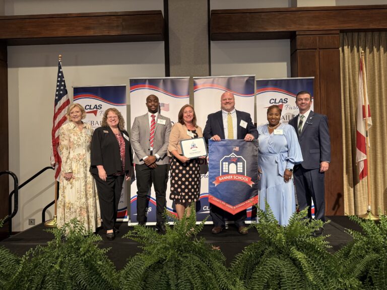 Group of seven educators and school leaders stand on stage at the CLAS Banner School Awards ceremony, posing with a Banner School banner and award plaque. American and Alabama flags flank the stage, with CLAS sponsor backdrops and greenery in front of the platform.