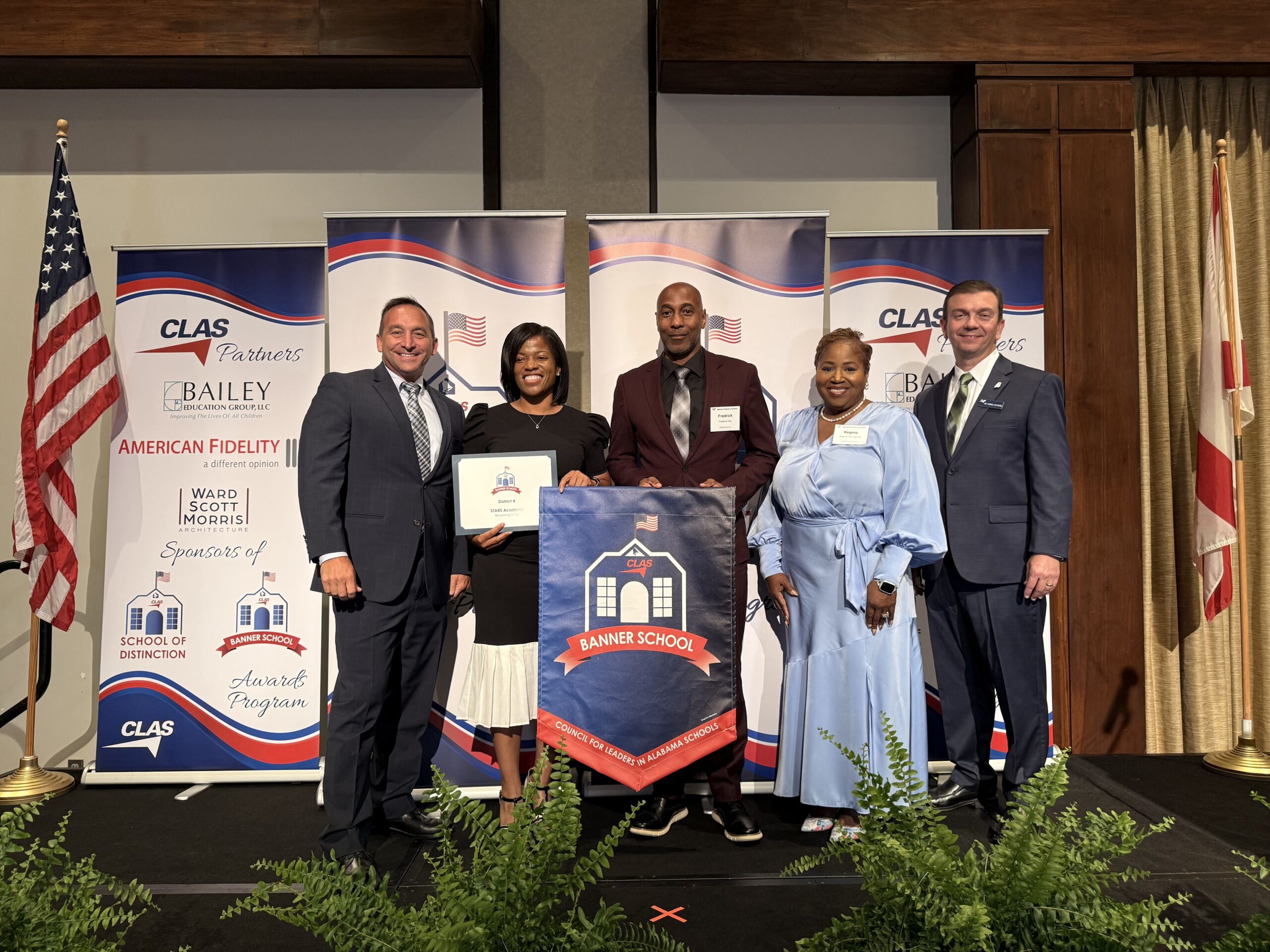Group of six educators and school leaders stand on stage at the CLAS Banner School Awards ceremony, posing with a Banner School banner and award plaque. American and Alabama flags frame the stage, with CLAS sponsor backdrops behind them and green ferns lining the front of the platform.
