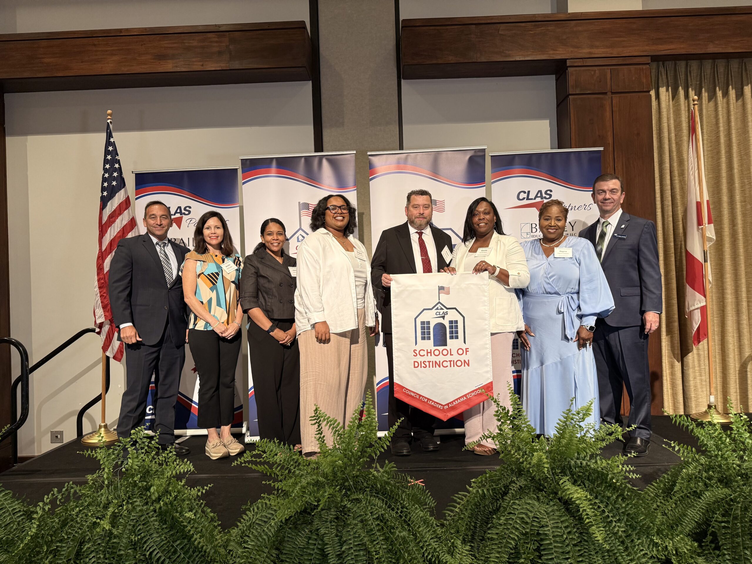 Group of nine educators and school leaders stand on stage at the CLAS School of Distinction Awards ceremony, posing with a School of Distinction banner. American and Alabama flags frame the stage, with CLAS sponsor backdrops behind them and green ferns lining the front of the platform.