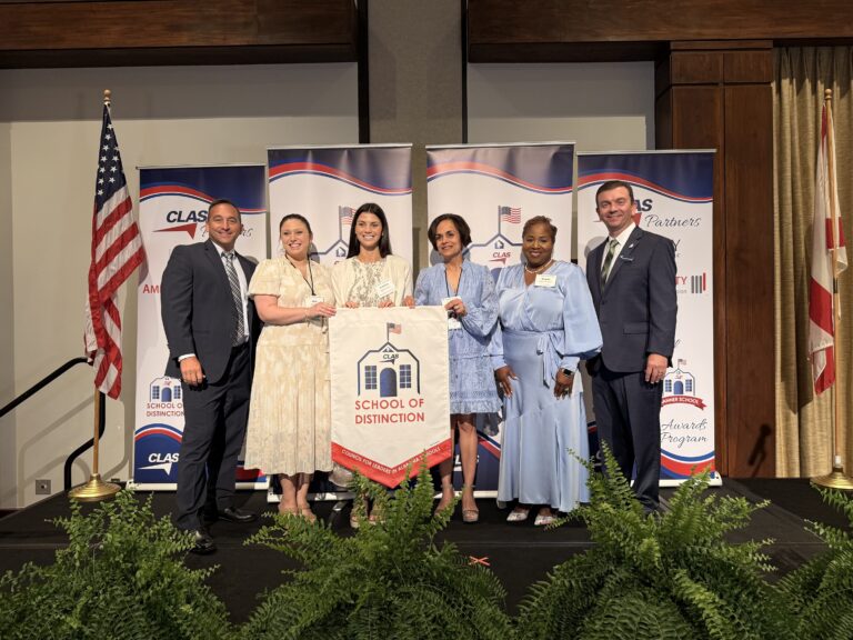 Group of six educators and school leaders stand on stage at the CLAS School of Distinction Awards ceremony, posing with a School of Distinction banner. American and Alabama flags frame the stage, with CLAS sponsor backdrops behind them and green ferns lining the front of the platform.