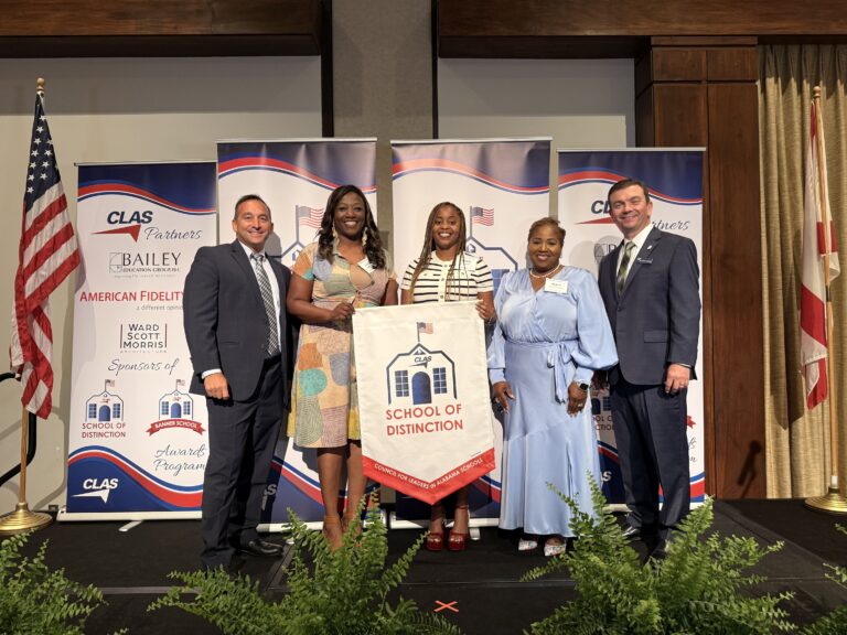 Group of five educators and school leaders stand on stage at the CLAS School of Distinction Awards ceremony, posing with a School of Distinction banner. American and Alabama flags frame the stage, with CLAS sponsor backdrops behind them and green ferns lining the front of the platform.