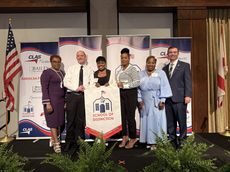 Group of seven educators and school leaders stand on stage at the CLAS School of Distinction Awards ceremony, posing with a School of Distinction banner. American and Alabama flags frame the stage, with CLAS sponsor backdrops behind them and green ferns lining the front of the platform.