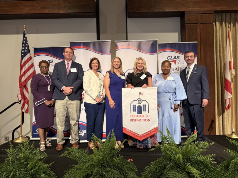 Group of eight educators and school leaders stand on stage at the CLAS School of Distinction Awards ceremony, posing with a School of Distinction banner. American and Alabama flags frame the stage, with CLAS sponsor backdrops behind them and green ferns lining the front of the platform.