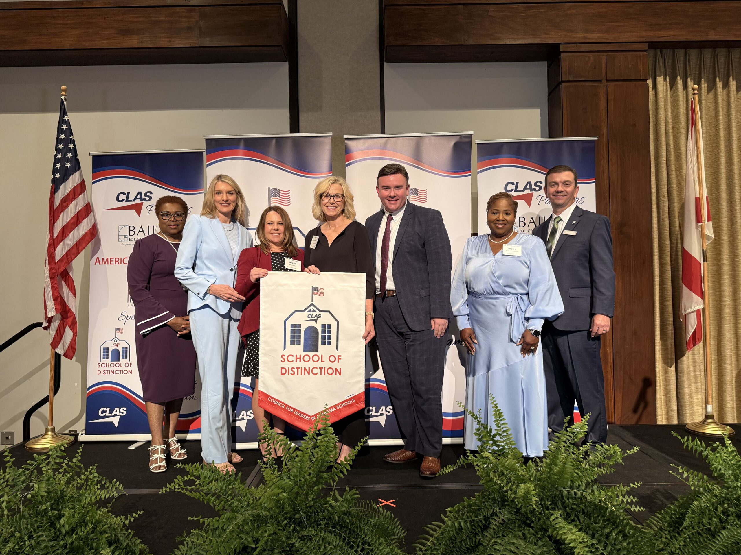 Group of eight educators and school leaders stand on stage at the CLAS School of Distinction Awards ceremony, posing with a School of Distinction banner. American and Alabama flags frame the stage, with CLAS sponsor backdrops behind them and green ferns lining the front of the platform.