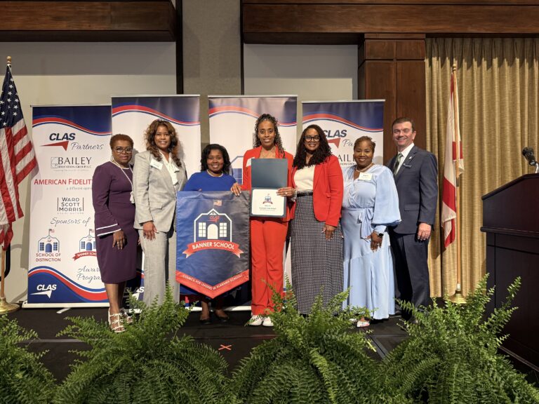 Group of eight educators and school leaders stand on stage at the CLAS Banner School Awards ceremony, posing with a Banner School banner and an award plaque. American and Alabama flags frame the stage, with CLAS sponsor backdrops behind them and green ferns lining the front of the platform.
