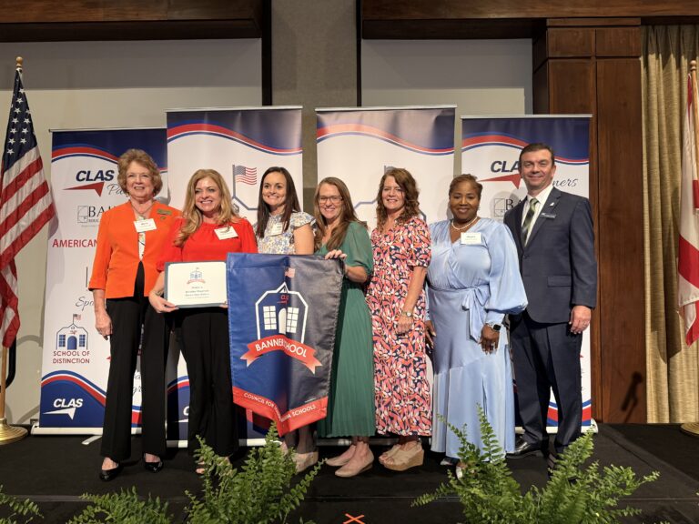 Group of eight educators and school leaders stand on stage at the CLAS School of Distinction Awards ceremony, posing with a School of Distinction banner. American and Alabama flags frame the stage, with CLAS sponsor backdrops behind them and green ferns lining the front of the platform.