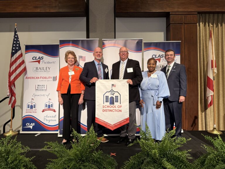 Group of six educators and school leaders stand on stage at the CLAS School of Distinction Awards ceremony, posing with a School of Distinction banner. American and Alabama flags frame the stage, with CLAS sponsor backdrops behind them and green ferns lining the front of the platform.
