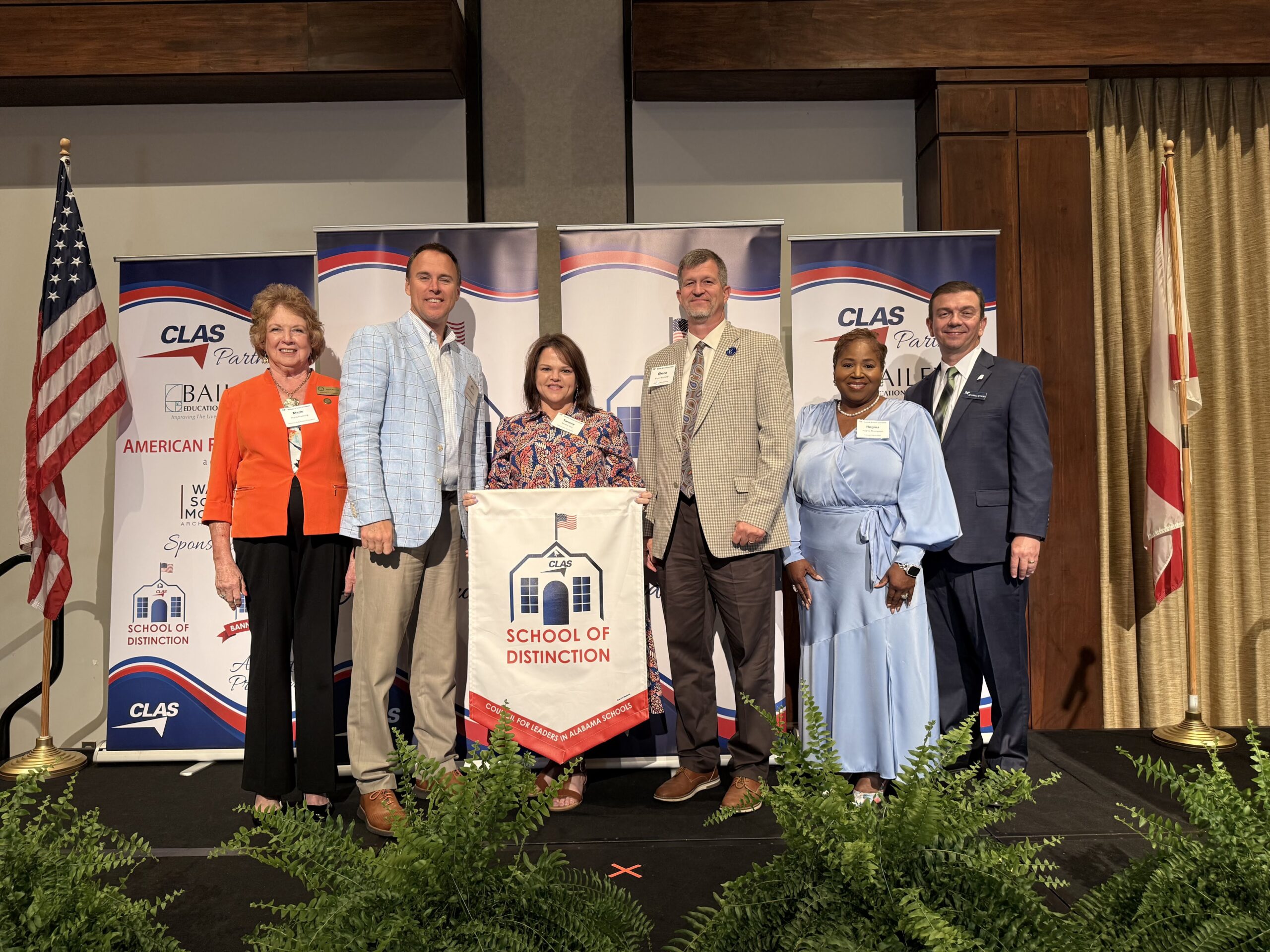 Group of six educators and school leaders stand on stage at the CLAS School of Distinction Awards ceremony, posing with a School of Distinction banner. American and Alabama flags frame the stage, with CLAS sponsor backdrops behind them and green ferns lining the front of the platform.