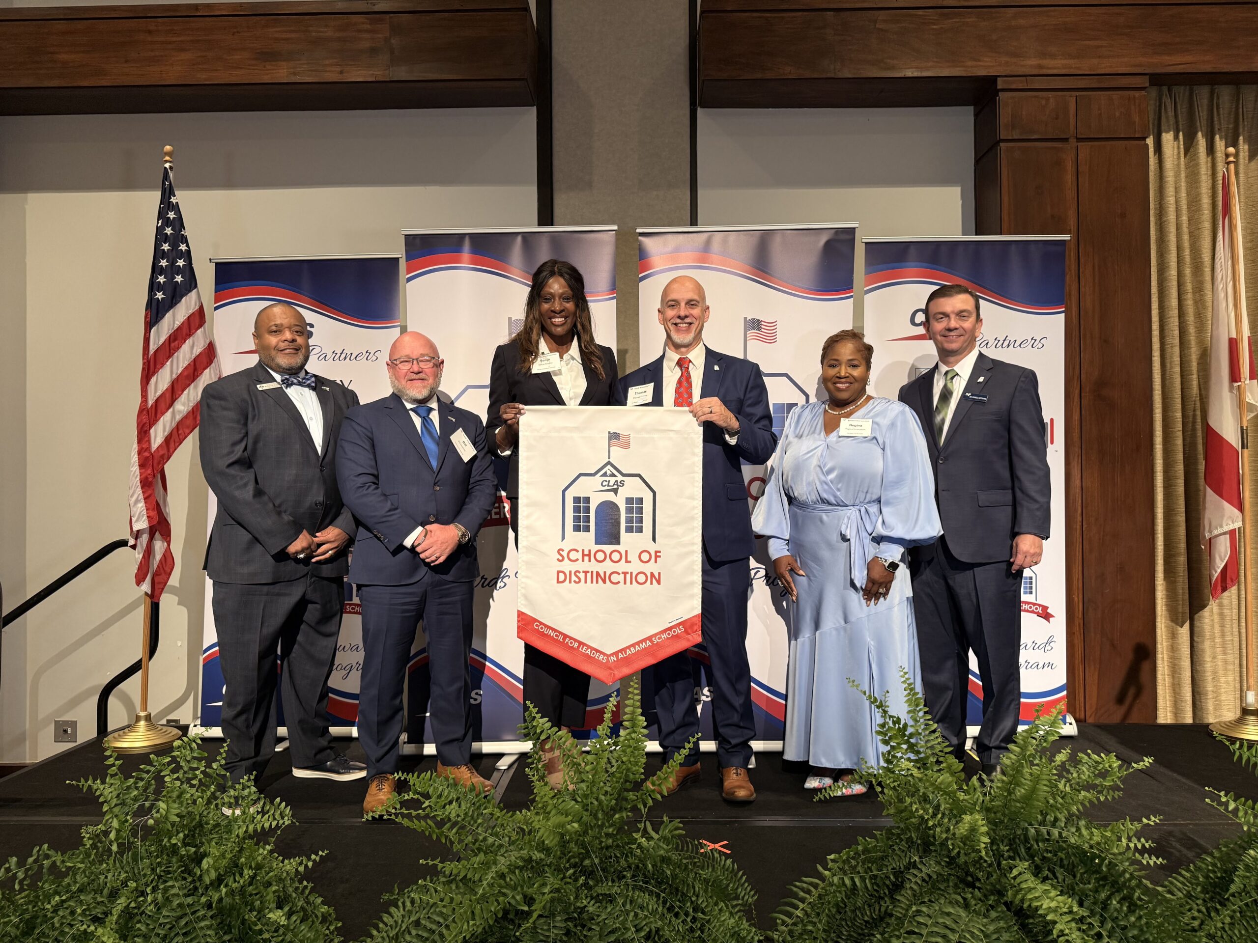 Group of seven educators and school leaders stand on stage at the CLAS School of Distinction Awards ceremony, posing with a School of Distinction banner. American and Alabama flags frame the stage, with CLAS sponsor backdrops behind them and green ferns lining the front of the platform.