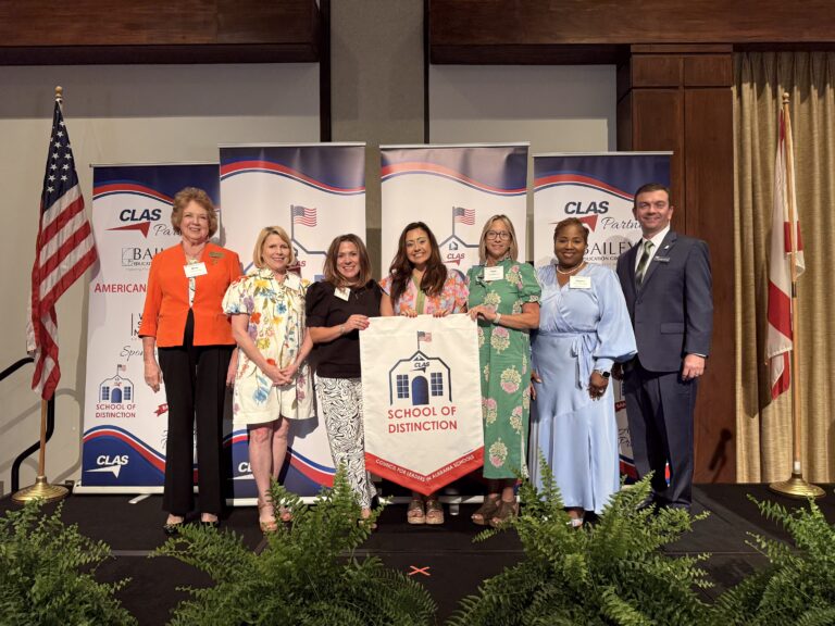 Group of seven educators and school leaders stand on stage at the CLAS School of Distinction Awards ceremony, posing with a School of Distinction banner. American and Alabama flags frame the stage, with CLAS sponsor backdrops behind them and green ferns lining the front of the platform.