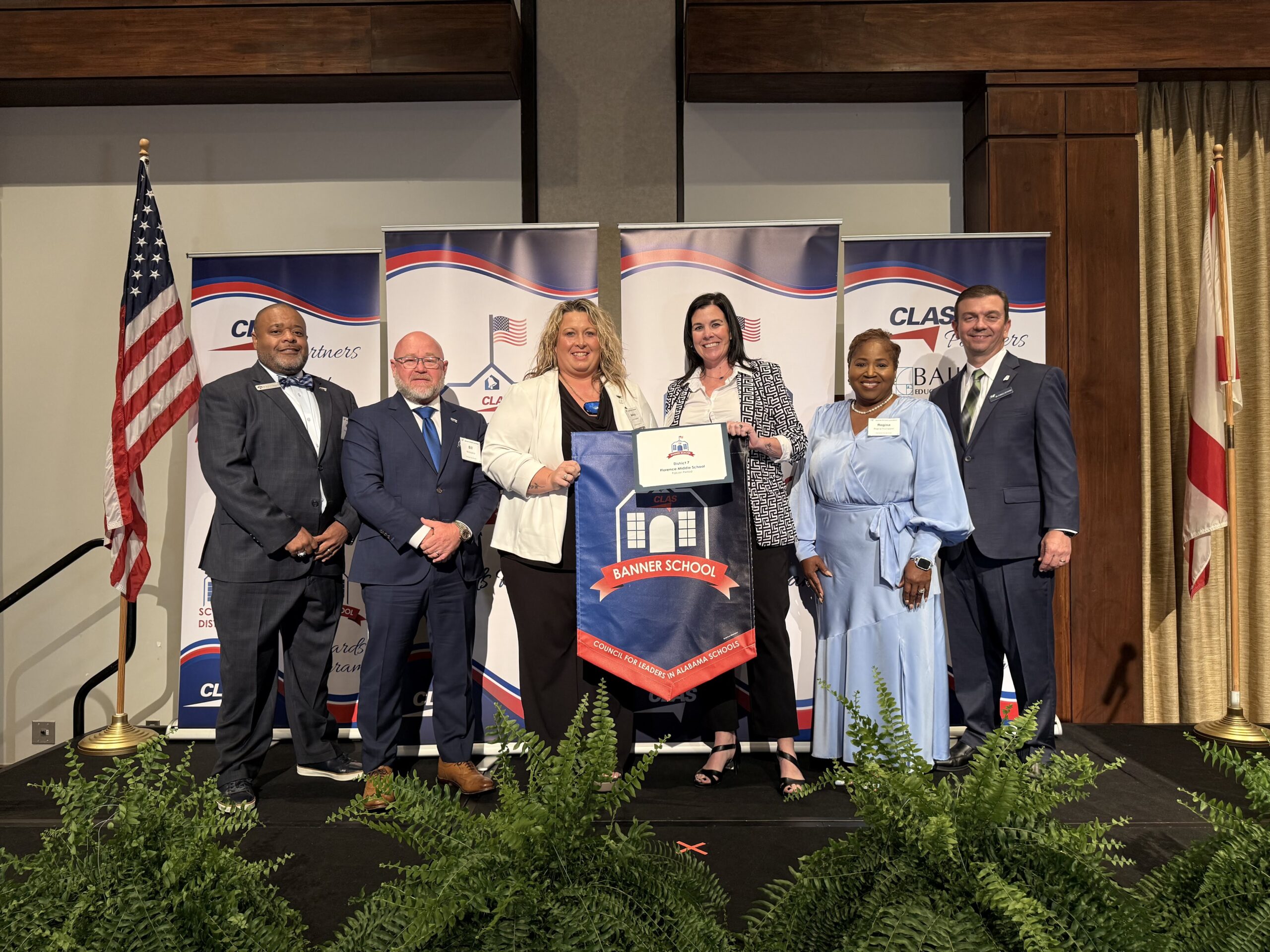 Group of seven educators and school leaders stand on stage at the CLAS Banner School Awards ceremony, posing with a Banner School banner and award plaque. American and Alabama flags frame the stage, with CLAS sponsor backdrops behind them and green ferns lining the front of the platform.