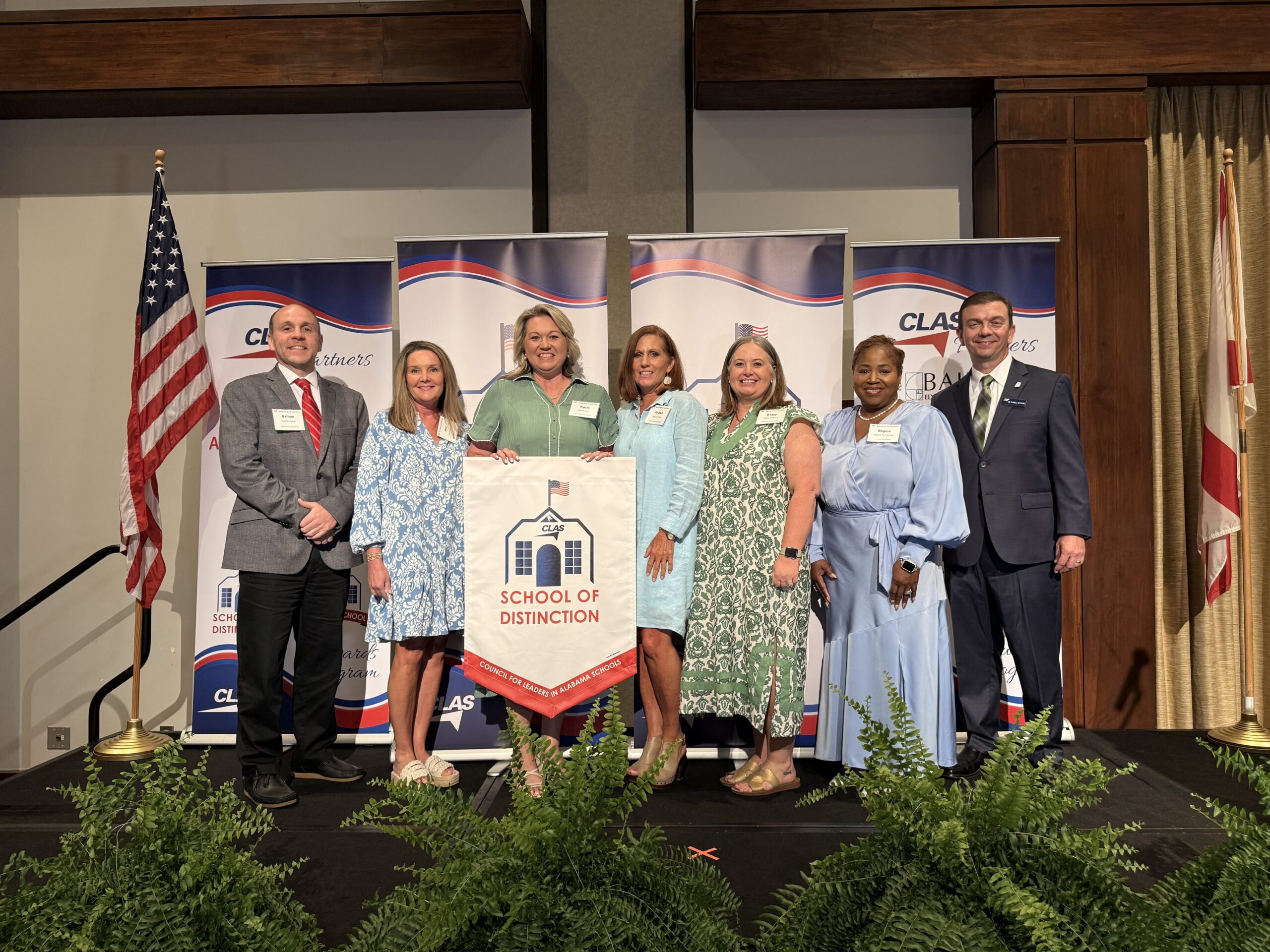 Group of seven educators and school leaders stand on stage at the CLAS School of Distinction Awards ceremony, posing with a School of Distinction banner. American and Alabama flags frame the stage, with CLAS sponsor backdrops behind them and green ferns lining the front of the platform.