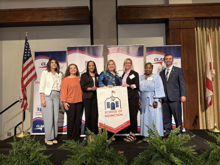 Group of seven educators and school leaders stand on stage at the CLAS School of Distinction Awards ceremony, posing with a School of Distinction banner. American and Alabama flags frame the stage, with CLAS sponsor backdrops behind them and green ferns lining the front of the platform.