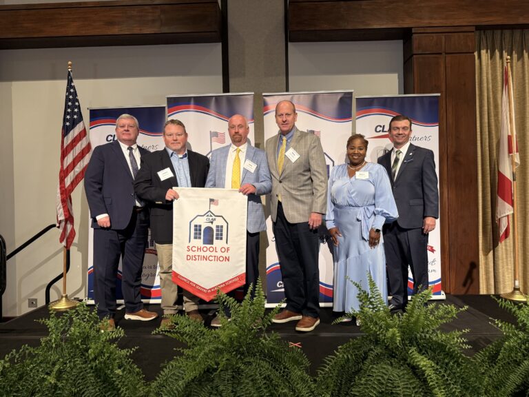 Seven educators and school leaders stand on stage at the CLAS Awards ceremony, posing with a “School of Distinction” banner. CLAS-branded sponsor backdrops are behind them, with the American and Alabama flags on either side and green ferns lining the front of the stage.