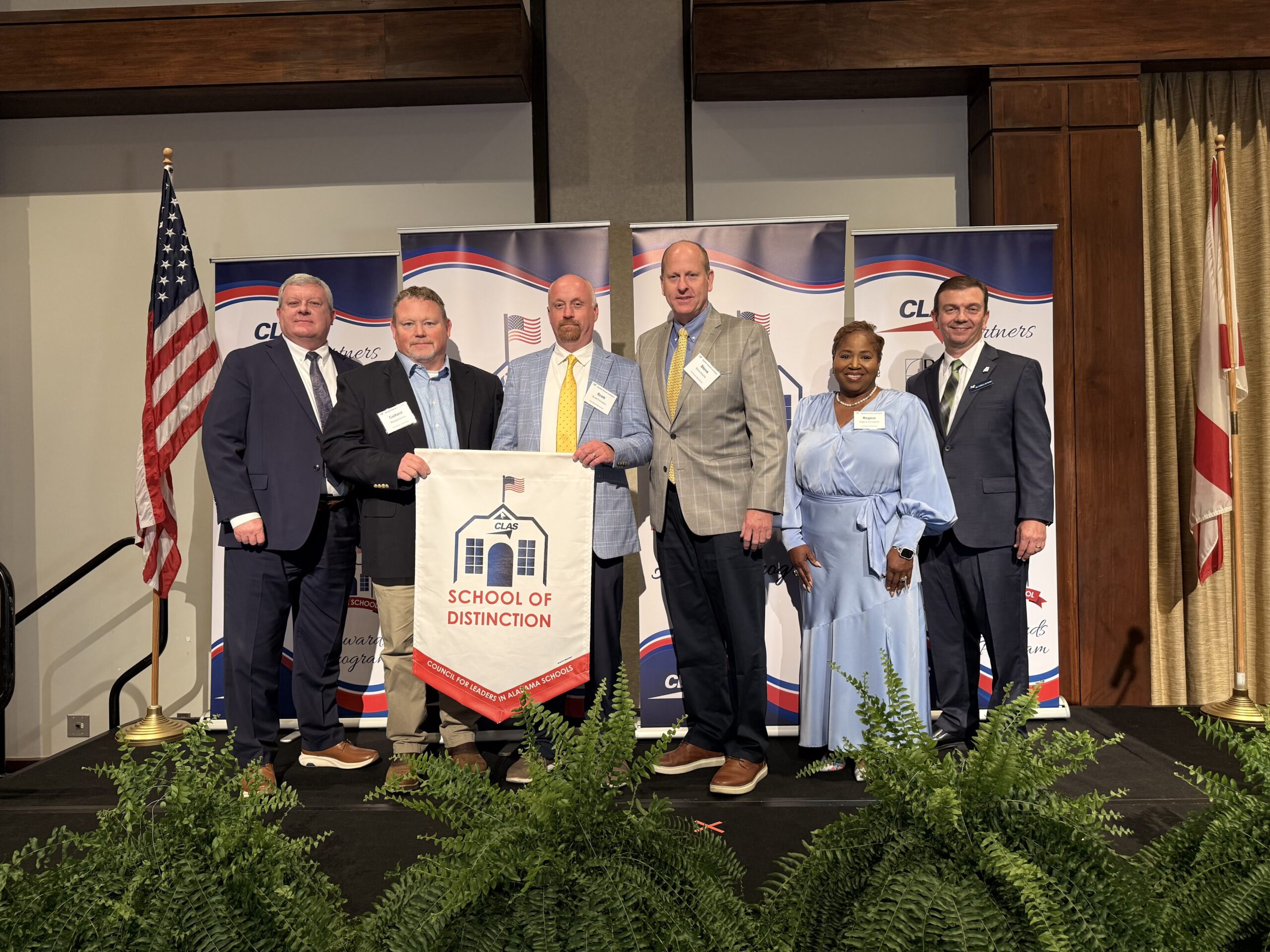 Seven educators and school leaders stand on stage at the CLAS Awards ceremony, posing with a “School of Distinction” banner. CLAS-branded sponsor backdrops are behind them, with the American and Alabama flags on either side and green ferns lining the front of the stage.