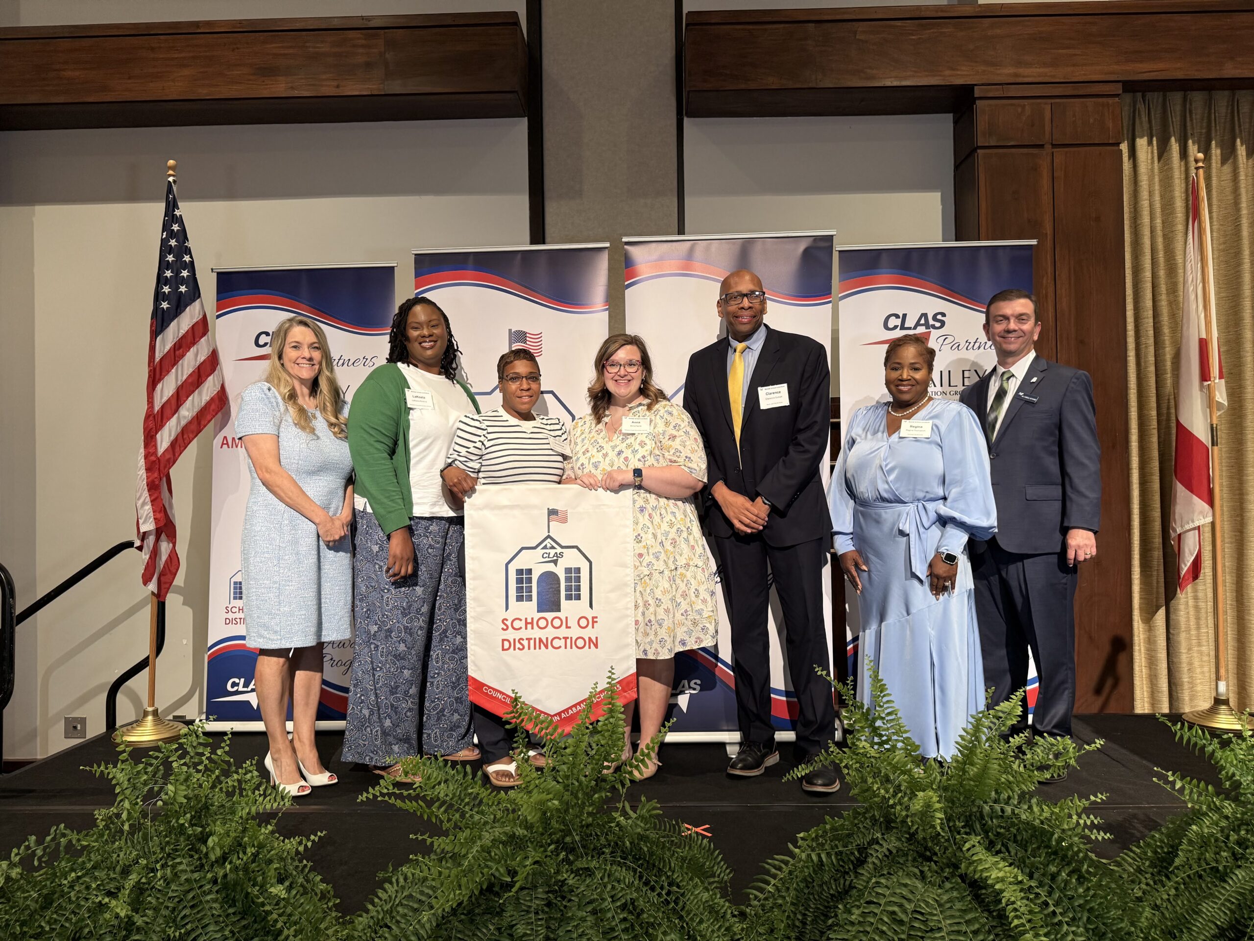 Eight educators and school leaders stand on stage at a CLAS awards ceremony, posing with a “School of Distinction” banner. CLAS-branded sponsor backdrops are behind them, with the American and Alabama flags on either side and green ferns lining the front of the stage.