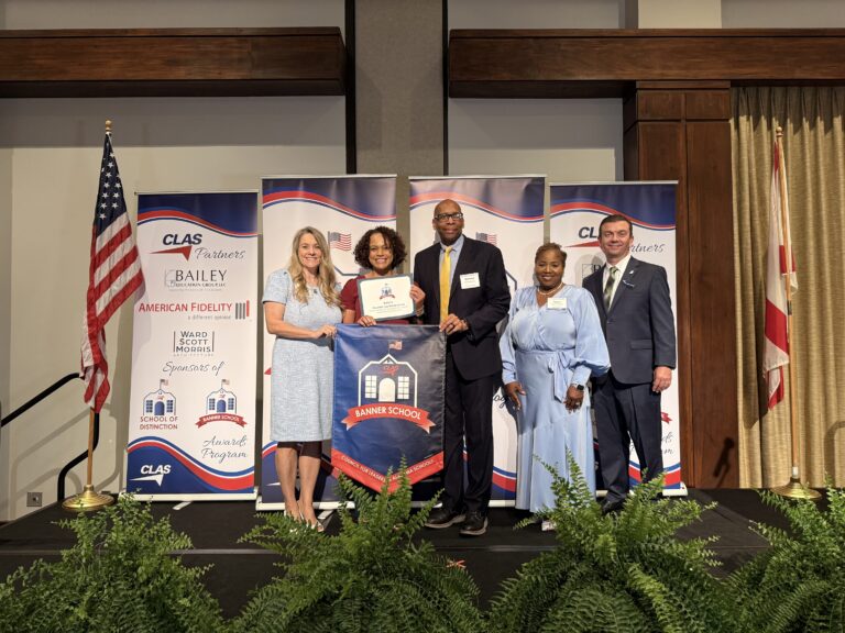 Five educators and school leaders stand on stage at a CLAS awards ceremony, posing with a “Banner School” banner and a certificate. CLAS-branded sponsor backdrops are behind them, with the American and Alabama flags on either side and green ferns lining the front of the stage.