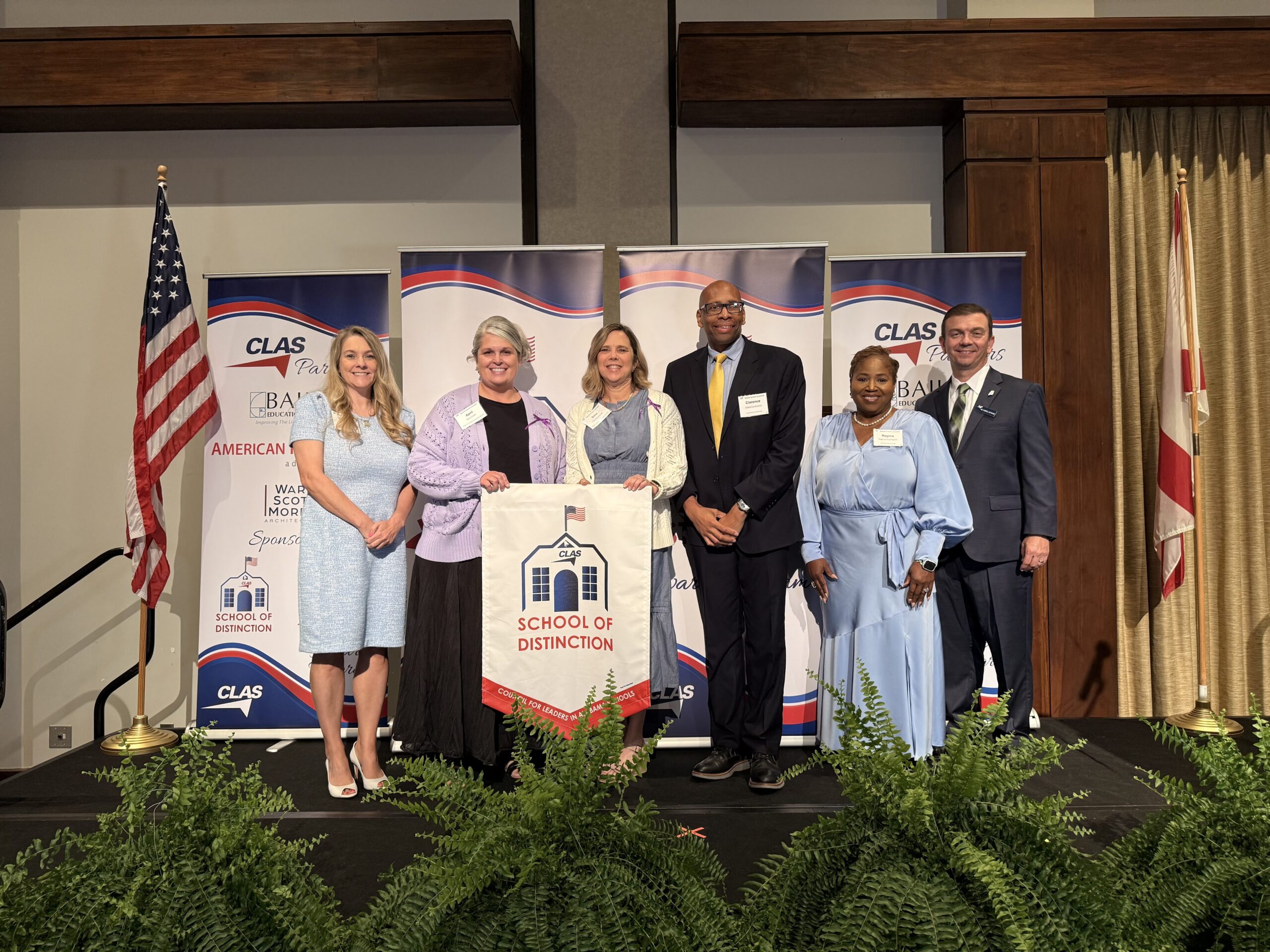 Seven educators and school leaders stand on stage at a CLAS awards ceremony, posing with a “School of Distinction” banner. CLAS-branded sponsor backdrops are behind them, with the American and Alabama flags on either side and green ferns lining the front of the stage.