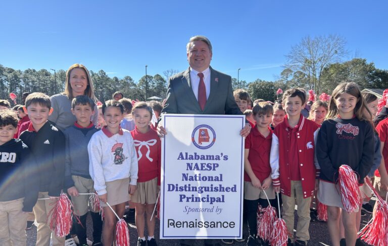 Stan Stokley, principal of Saraland Elementary School, stands with a group of smiling students holding red and white pom-poms. He holds a sign recognizing him as Alabama’s NAESP National Distinguished Principal, with a crowd of cheering students in the background.