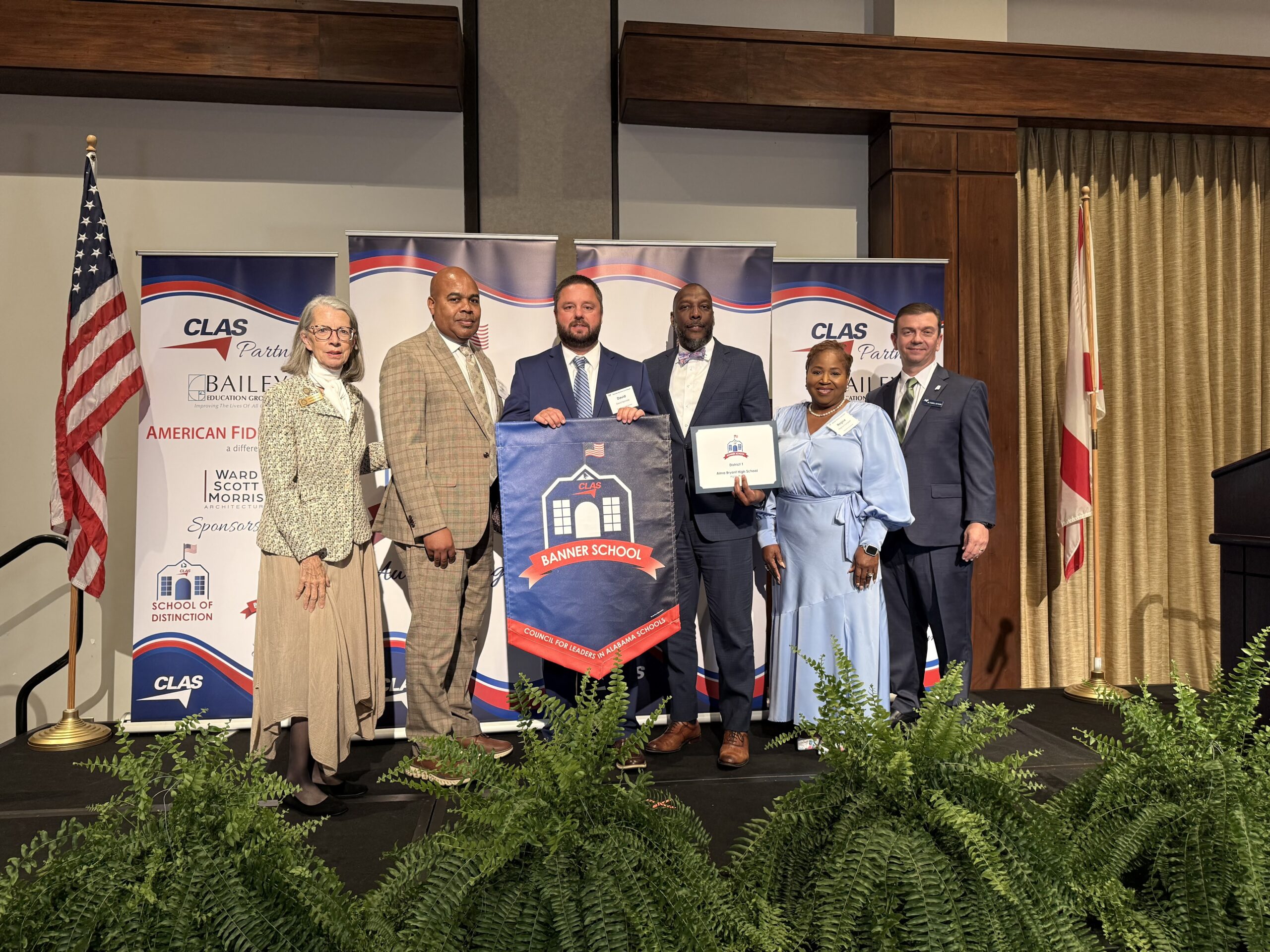 Group of seven educators and school leaders stand on stage at the CLAS Banner School Awards ceremony, posing with a Banner School banner and award plaque. American and Alabama flags frame the stage, with CLAS sponsor backdrops behind them and green ferns lining the front of the platform.